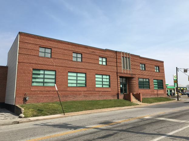 Ein großes Backsteinschulgebäude an einer Straßenecke mit Glasfenstern, Treppen, Geländern, Rasen, Laternen, Schilder, Fahrzeugen, Bäumen und einem bewölkten Himmel im Hintergrund.