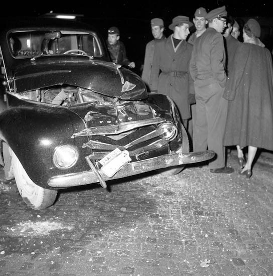 Schwarz-weißes Bild eines schwerbeschädigten Autos am Straßenrand mit eingedellter Motorhaube und zersplitterten Fenstern, umgeben von Menschen, die die Szene begutachten.