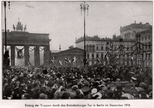 Eine große Menge steht vor dem Brandenburger Tor in Berlin, Deutschland im Jahr 1918, trägt Hüte und hält Fahnen, wobei die Säulen, Statuen und umliegenden Gebäude im Hintergrund zu sehen sind.