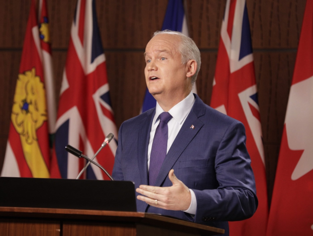 Ein Mann im Anzug und Krawatte steht an einem Rednerpult mit einem Laptop und zwei Mikrofonen und hält eine Rede mit Flaggen im Hintergrund.