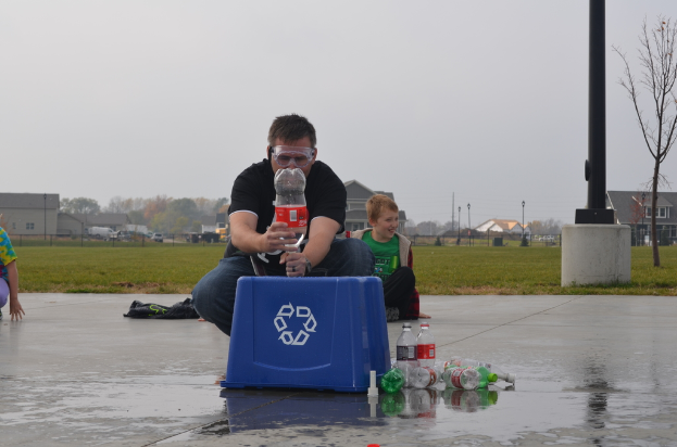 Ein Mann mit Brille hält eine Flasche, umgeben von ein paar weiteren Flaschen und einer Schachtel, mit Pfählen, Bäumen, Gebäuden, Fahrzeugen und Gras im Hintergrund.