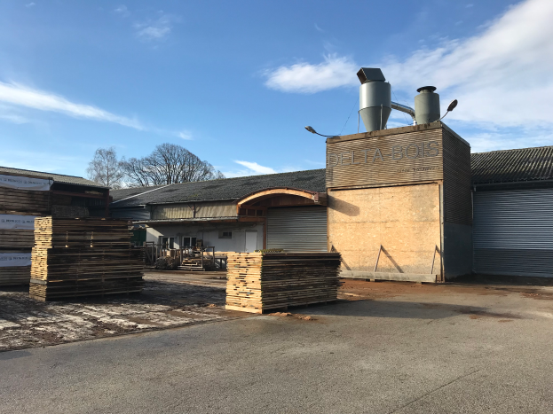Ein Gebäude mit der Aufschrift "Delta Bois Destillerie" und Holzplanken davor, umgeben von Bäumen unter einem klaren blauen Himmel.