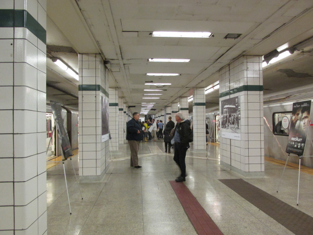Eine U-Bahn-Station mit Menschen, die gehen, Säulen, Deckenleuchten und einem wartenden Zug auf dem rechten Gleis.