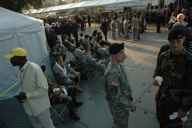 Eine Gruppe von Menschen in Militäruniformen versammelte sich draußen, wobei einige auf Stühlen saßen, Zelte im Hintergrund und ein Baum oben im Bild.