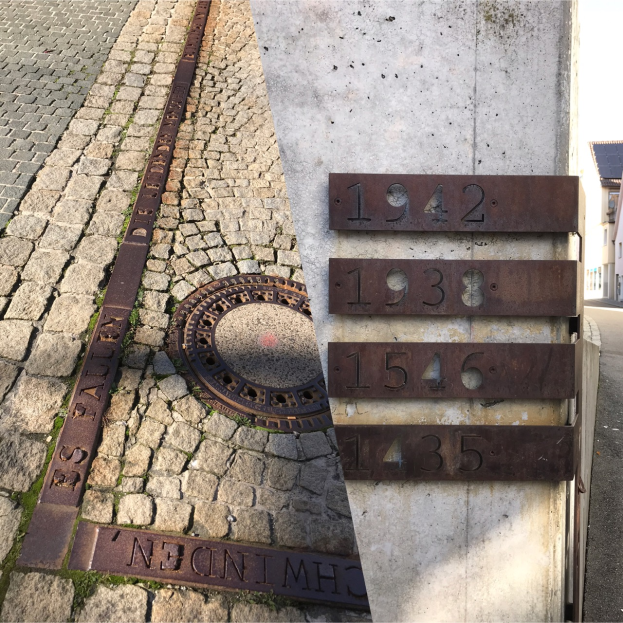 Eine Kopfsteinpflasterstraße mit einem Gullideckel, einer nummerierten Wand auf der rechten Seite und Gebäuden unter einem klaren blauen Himmel im Hintergrund.