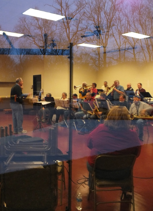 Musiker mit Trompeten im Hintergrund, Sänger vor Mikrofonen und ein Mann, der Musik komponiert, mit einer Wand und Bäumen im Glasreflexion sichtbar.