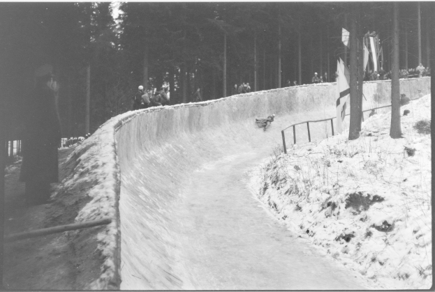 Ein Schwarz-Weiß-Foto eines Menschen, der auf einem Bobschlitten eine schneebedeckte Bahn hinunterfährt, umgeben von einem Zaun und Bäumen, mit ein paar Menschen im Hintergrund.