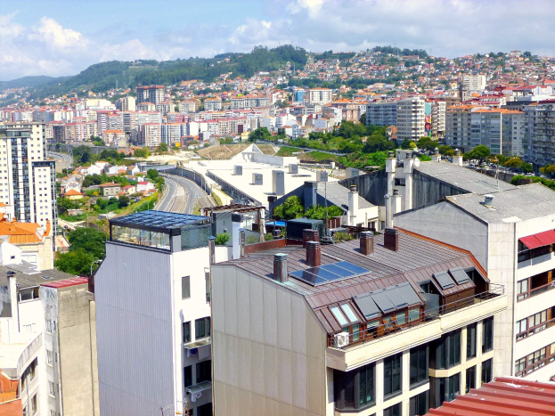 Eine Stadtansicht von einem hohen Standpunkt aus, die zahlreiche Gebäude, Bäume, Straßen, Fahrzeuge und entfernte Berge unter einem bewölkten Himmel zeigt, wobei auf dem Dach eines Gebäudes Solarpaneele zu sehen sind.