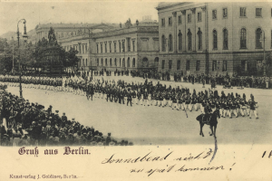 Ein altes Schwarz-Weiß-Foto einer Parade in Berlin, Deutschland, mit Menschen auf dem Boden und auf Pferden, einer Statue auf einem Sockel, Straßenlaternen, Bäumen, Gebäuden mit Fenstern und einem bewölkten Himmel, mit Text am unteren Rand des Bildes.
