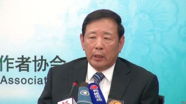 Ein Mann im Anzug und Krawatte sitzt an einem Tisch mit Mikrofonen und spricht vor einem Banner, wahrscheinlich zu Nachrichten über Chinas Präsident Hu Jintao.