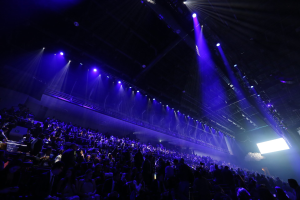 Eine große Menschenmenge sitzt einer Bühne mit Deckenbeleuchtung gegenüber, und auf der rechten Seite ist ein Bildschirm zu sehen, was auf eine Konzerthalle hinweist.