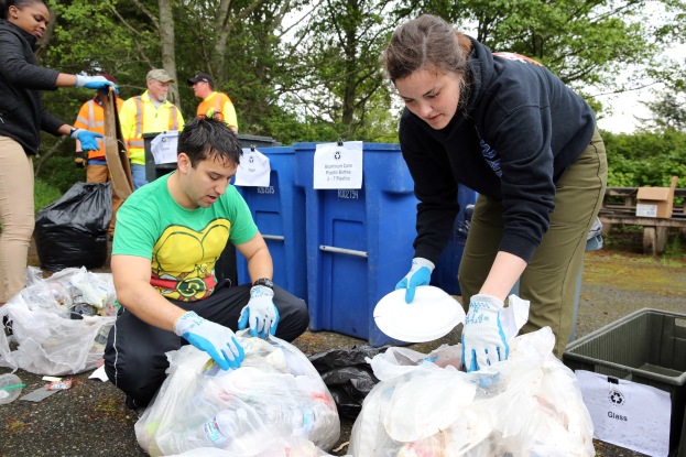 Eine Gruppe von Menschen, die Müll in einem Park sammeln, mit einem Mann und einer Frau in Handschuhen, die Teller halten, umgeben von Plastikmüll, Flaschen und Schutt, neben einem Mülleimer und einer Holzbank, mit Bäumen und einem klaren blauen Himmel im Hintergrund.