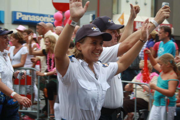 Eine Gruppe von Menschen, einige mit Mützen und Dosen, stehen und sitzen vor einer Menge bei einem Umzug, mit Geländern, Luftballons und einem Gebäude mit Namensschildern im Hintergrund.