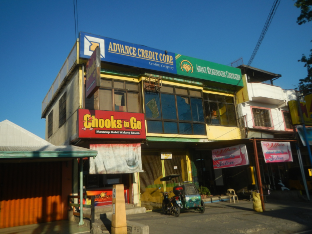 Außenansicht eines Gebäudes mit einer Tafel, auf der "Advance Credit Corp" steht, umgeben von Fahrzeugen, Strommasten und Bäumen unter einem klaren blauen Himmel.