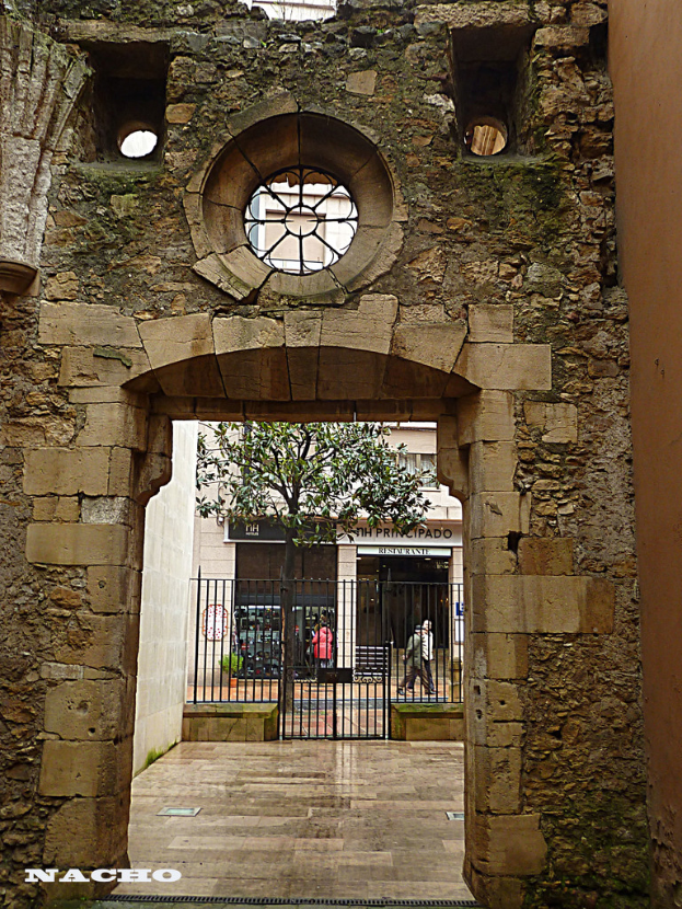 Ein eisernes Tor mit einem Bogen, das vor einem Gebäude steht, mit zwei Personen dahinter und einem Baum mit Zweigen und Blättern zu sehen, mit einem Wasserzeichen am unteren Bildrand.