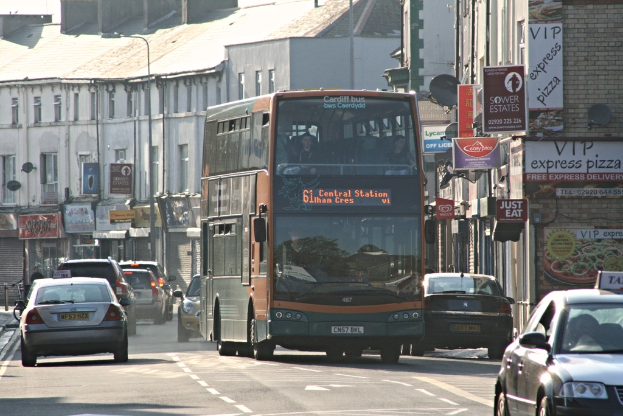 Eine Straße mit Autos und einem Bus vor Gebäuden mit Wänden, Fenstern, Tellern und Dächern, die Plakate und Banner an den Wänden und einen Mast mit einer Straßenlaterne zeigen.