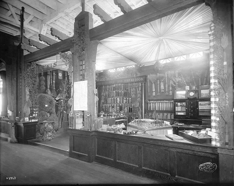 Altes Schwarz-Weiß-Foto eines Apothekeninneren mit einem zentralen Tresen, auf dem verschiedene Gegenstände und im Hintergrund Regale mit Flaschen und Gegenständen stehen.