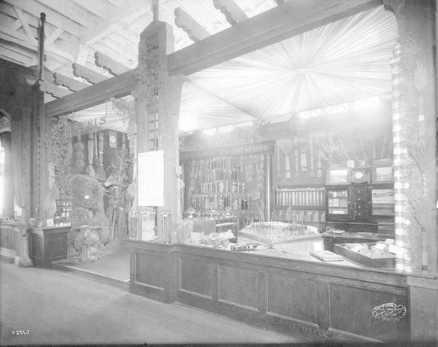 Altes Schwarz-Weiß-Foto eines Apothekeninneren mit einem zentralen Tresen, auf dem verschiedene Gegenstände und im Hintergrund Regale mit Flaschen und Gegenständen stehen.