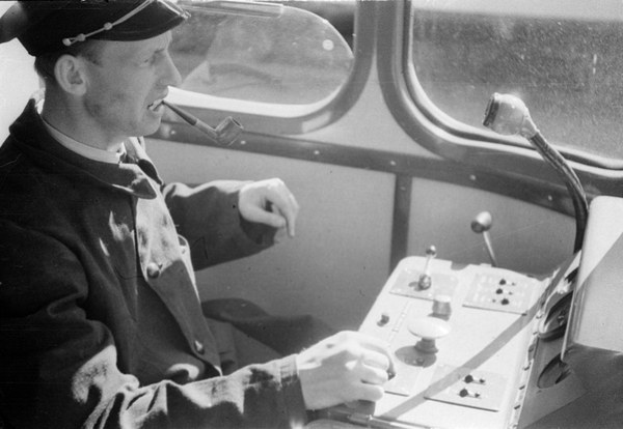 Schwarz-weißes Bild eines Mannes in Mütze und Uniform, der an den Kontrollen eines Zuges sitzt und eine Pfeife raucht, mit sichtbaren Glasfenstern im Hintergrund.