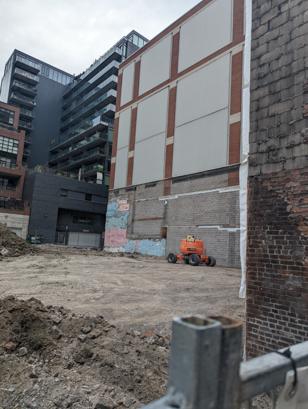 Baustelle mit einem teilweise abgerissenen Gebäude im Hintergrund, einem Fahrzeug am Boden, einem Metallzaun und einem bewölkten Himmel.