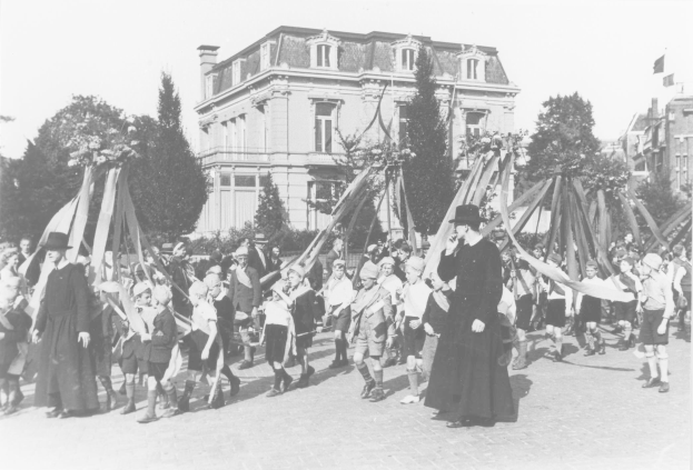 Schwarze und weiße Aufnahme einer Gruppe von Menschen, die eine Straße entlanggehen und Fahnen schwenken, mit Hüten, Bäumen, Gebäuden und einem Maibaum im Hintergrund.