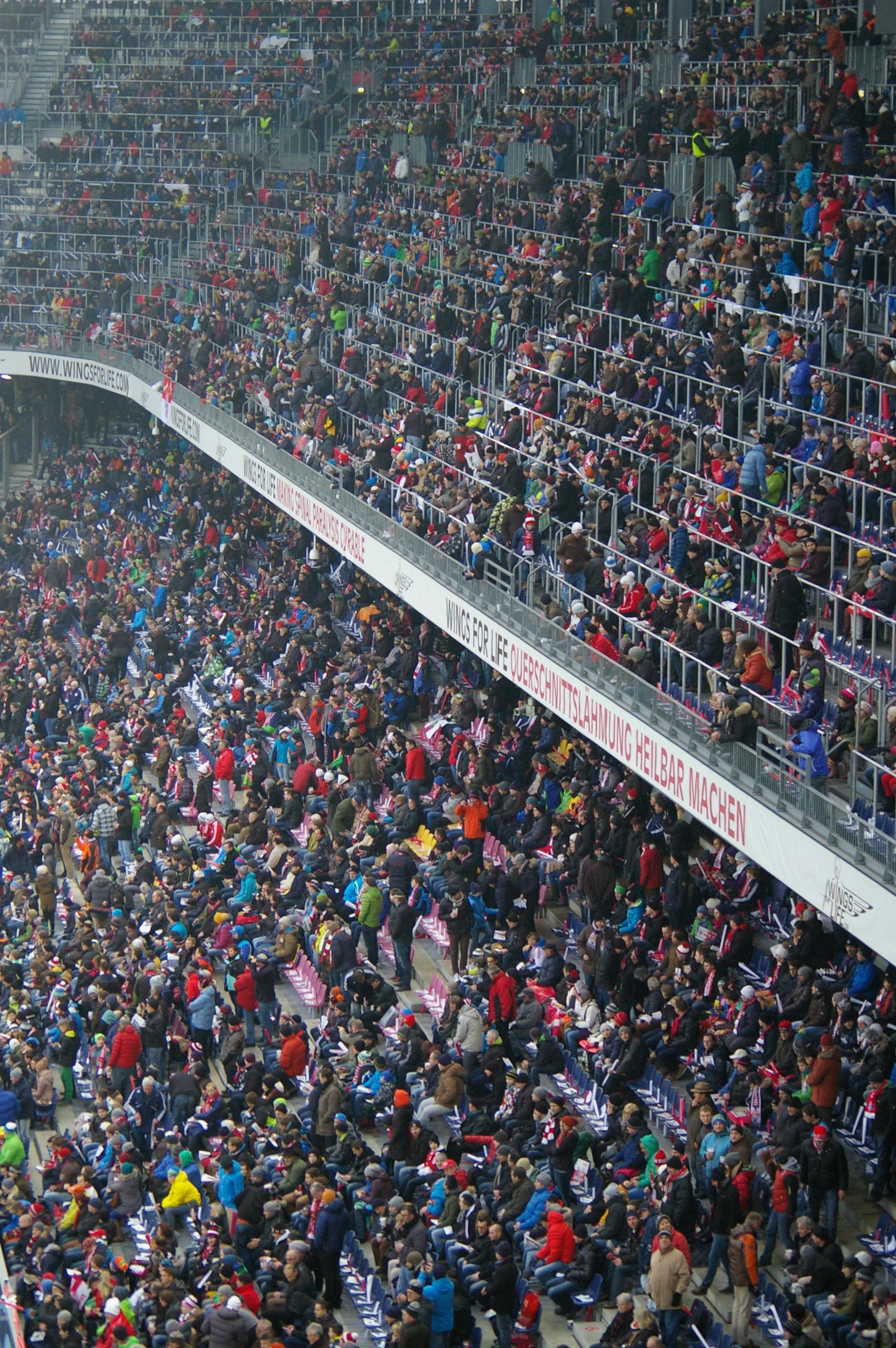 Eine große Menschenmenge sitzt und steht in einem Stadion mit Geländern und Texttafeln im Hintergrund.