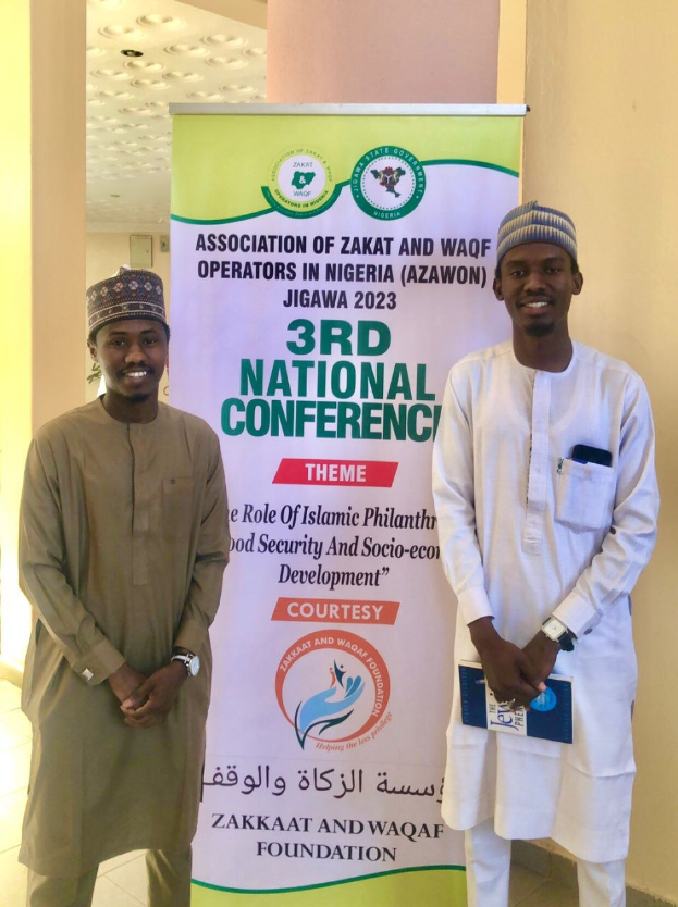 Zwei Männer stehen nebeneinander vor einem Banner mit der Aufschrift "3. Nationalkonferenz" mit einer Säule und Deckenlampen im Hintergrund.