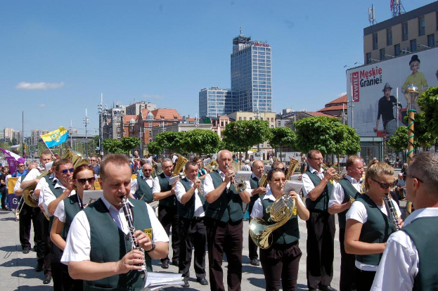 Eine Gruppe von Menschen in passenden Kostümen, die verschiedene Musikinstrumente spielen, draußen mit Bäumen, Pfählen und einem Banner mit Text und Bildern im Hintergrund.