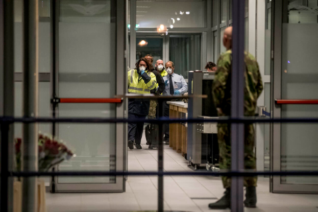 Eine Gruppe von Menschen mit Mund-Nasen-Schutz steht vor einer Glastür an einem Flughafen, mit einem Geländer im Vordergrund und einem Blumenstrauß auf der linken Seite.