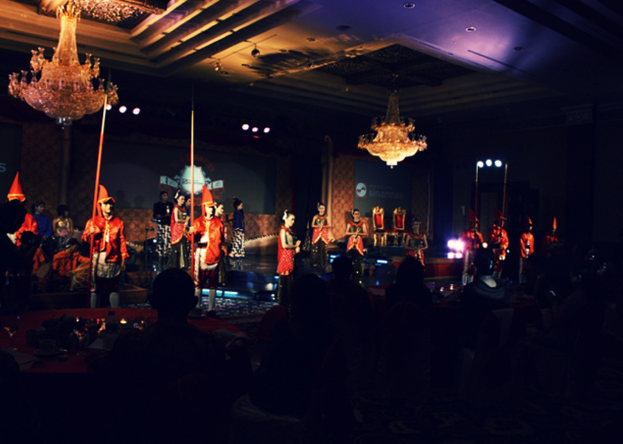 Eine Gruppe von Menschen sitzt und schaut einer Vorstellung zu, mit zwei Kronleuchtern an der Decke.