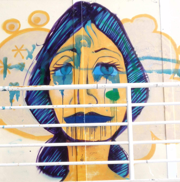 Ein Wandgemälde eines traurigen Frauenkopfs an einem Gebäude in Berlin, mit einem Gitter im Vordergrund, in gedämpften Farben gemalt.