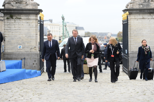 Eine Gruppe von Mitarbeitern geht in einen Bereich mit einem Tor, hinter dem sich viele Fahrzeuge und Gebäude befinden; eine Frau rechts trägt zwei Koffer.