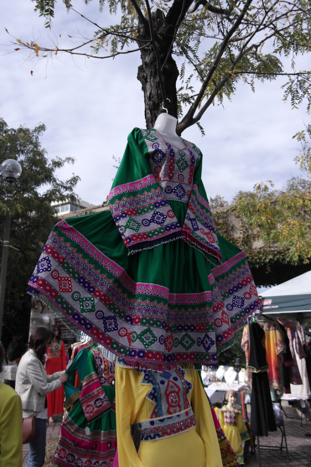 Eine Schaufensterpuppe steht in der Mitte eines belebten Einkaufsbereichs mit mehreren Geschäften, einer Person, die Kleider an einem Ständer arrangiert, einem Gebäude und Bäumen im Hintergrund.