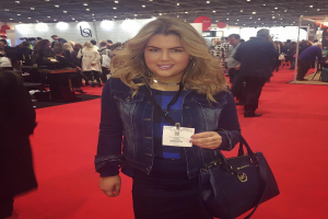 Eine Frau steht auf einem roten Teppich, lächelt und hält eine Handtasche in der Hand, mit Menschen, Lichtern und Tafeln im Hintergrund bei einer Modenschau.