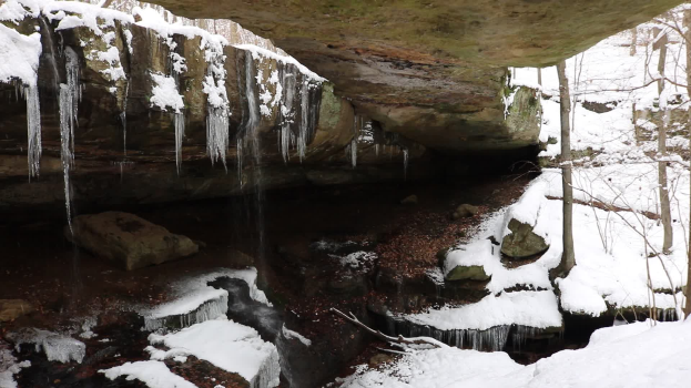 Ein kleiner Wasserfall ergießt sich über einen schneebedeckten, felsigen Hang in einem bewaldeten Gebiet, mit Eiszapfen an den Felsen.