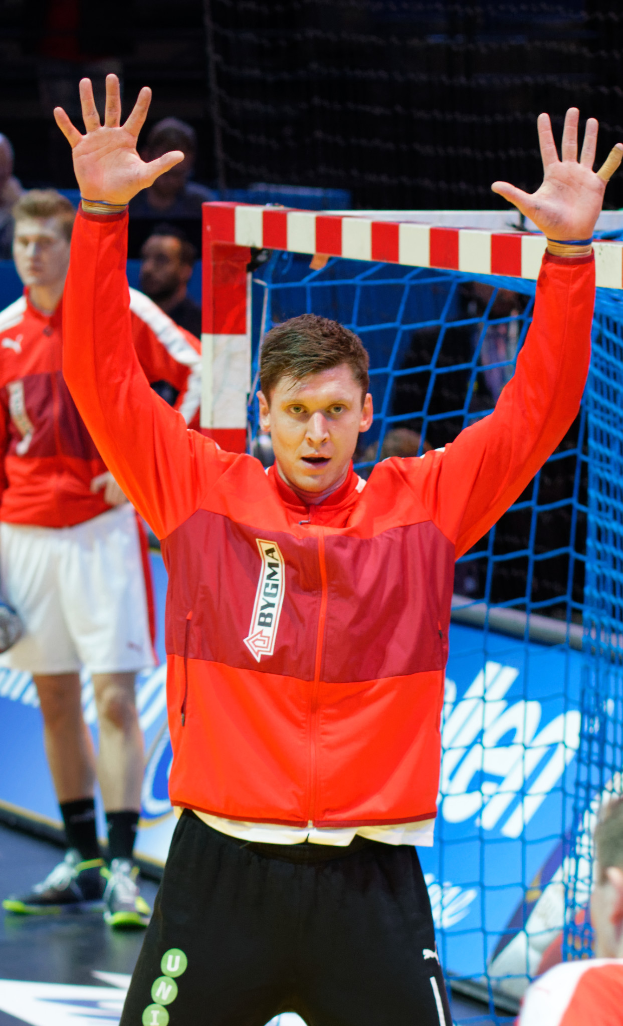 Ein Mann in einer roten Jacke und schwarzen Shorts steht auf einem Futsalplatz, trägt Schuhe und hat ein Netz und einige andere Personen im Hintergrund.