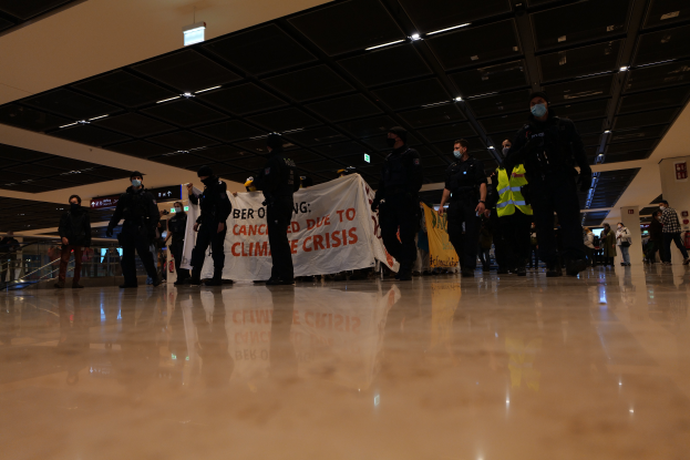 Eine Gruppe maskierter Polizisten in einem Flughafen, die ein Banner halten, umgeben von Gelöndern und Deckenlampen, mit Texttafeln im Hintergrund.