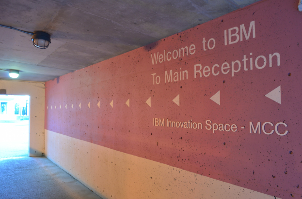 Eine Wand mit der Aufschrift "Willkommen bei IBM", beleuchtet von Deckenlampen, mit einer Tür im Hintergrund, die "IBM Innovation Space MCC" markiert.