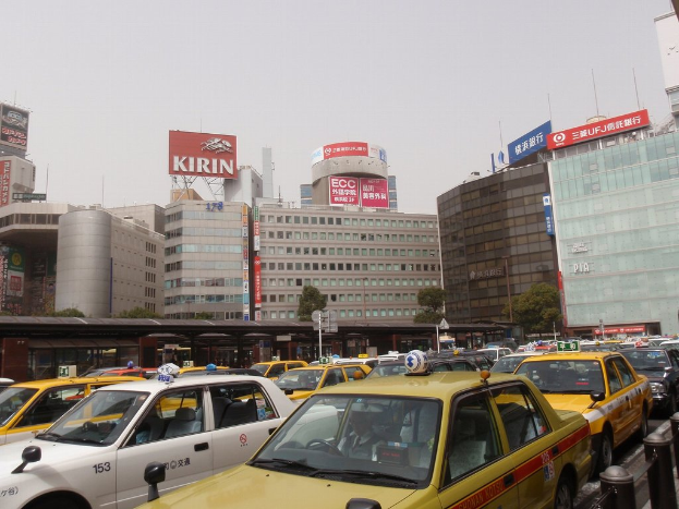 Eine Stadtansicht mit Fahrzeugen auf einer Straße, Gebäuden, Bäumen, Pfählen mit Schildern und Text auf den Schildern, mit einem Zug im Hintergrund und einem Geländer unten rechts.