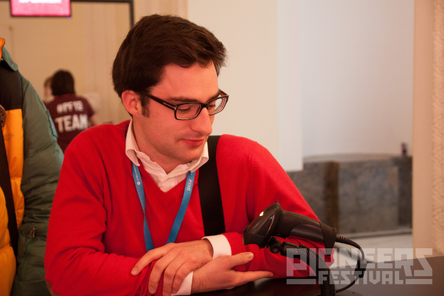 Ein Mann mit einer Ausweiskarte steht vor einem Mikrofon mit ein paar Menschen dahinter, eine Wand im Hintergrund, eine Tafel oben links und Text unten rechts.