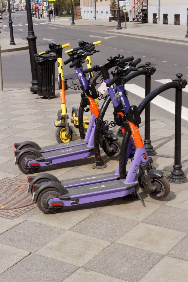 Eine Reihe geparkter Elektrokleinstfahrzeuge auf einer Stadtstraße mit einem Müllcontainer im Vordergrund und Gebäuden, Bäumen und Fahrzeugen im Hintergrund.