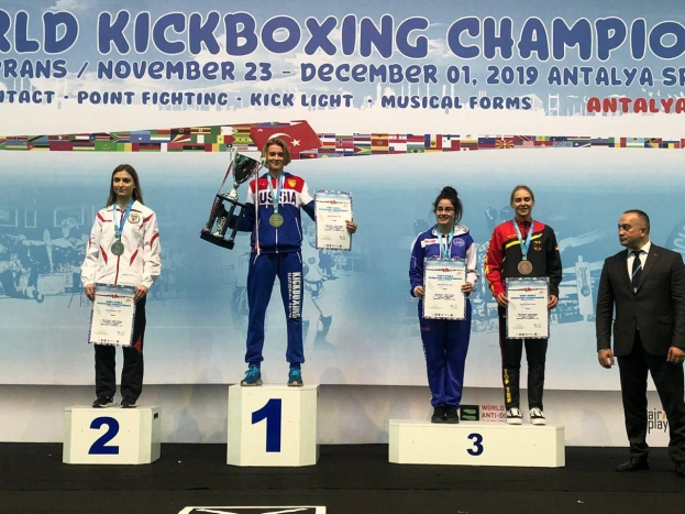 Fünf Personen auf einem Podium mit Troph├Ąen und Zertifikaten, im Hintergrund ein Banner 'World Kickboxing Championship Antalya 2019'.
