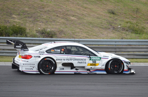 BMW M4 DTM-Rennwagen auf einer Rennstrecke mit Werbetext, umgeben von grünem Gras und einer Begrenzung im Hintergrund.