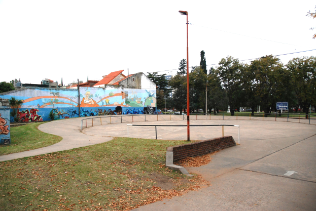 Eine Skate-Anlage mit einem Wandgemälde auf einem Gebäude, umgeben von Gras, trockenen Blättern, Laternen, Geländern und einer bemalten Wand, vor einem Hintergrund aus Bäumen, Gebäuden, Fahrzeugen und Himmel.