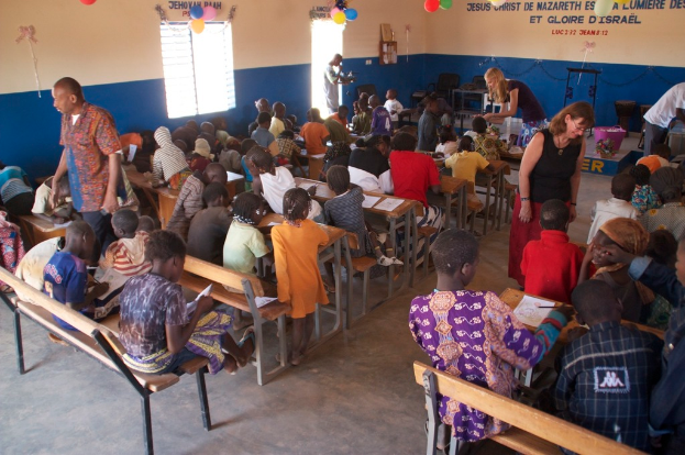 Kinder auf Bänken im Vordergrund, mit Erwachsenen, die sie führen, und einem Hintergrund mit Stühlen, einer Tür, einem Fenster, einem Tisch, Ballons und Text oben.