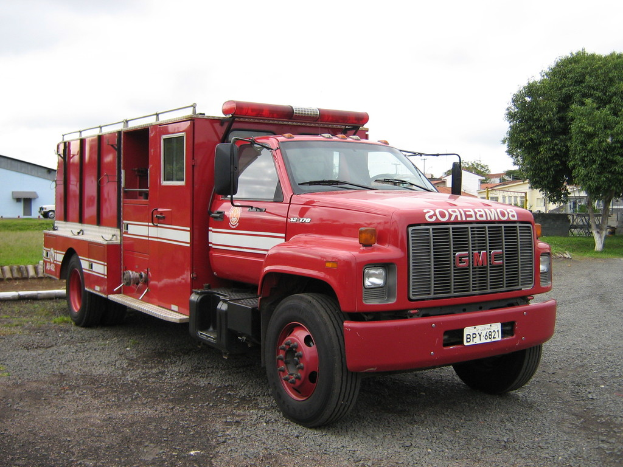 Motorisierte Fahrzeuge auf einer Straße mit Bäumen, Gras, Gebäuden und einem bewölkten Himmel im Hintergrund.