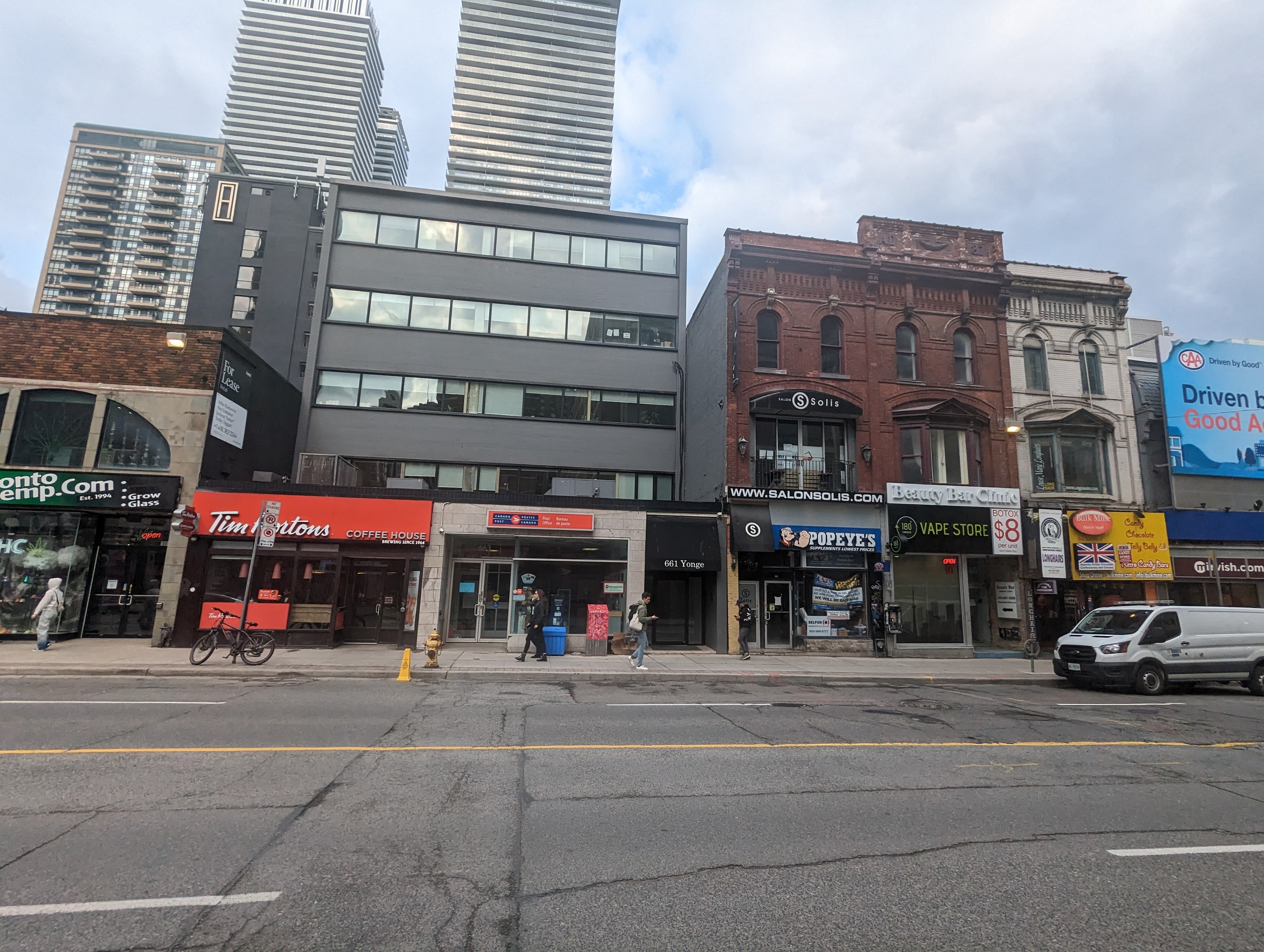 Stadtstraße mit hohen Gebäuden, fahrenden Fahrzeugen, Passanten, einem Fahrrad, einem Feuerhydranten und Vermietungsschildern unter einem bewölktem Himmel.
