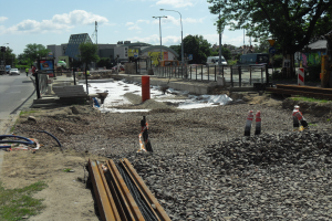 Eine Baustelle mit verschiedenen Geräten wie Steinen, Stangen, Verkehrszeichen, Rohren und Fahrzeugen, Gebäuden, Laternen, Bäumen und einem bewölkten Himmel im Hintergrund.