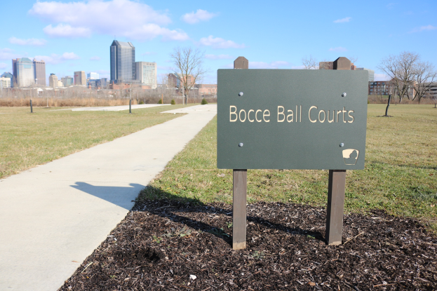 Ein Schild am Boden, das Boccia-Plätze in Indianapolis, Indiana, anzeigt, umgeben von Gras, Bäumen, einem Weg, Pfosten, einem Zaun, Gebäuden und einem bewölkten Himmel.
