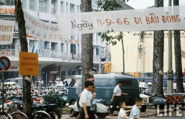 Menschen, Fahrräder, ein Van und Autos sind unten auf dem Bild zu sehen, im Hintergrund Gebäude und Bäume, sowie Schilder und Tafeln und ein Schild an der linken Seite.
